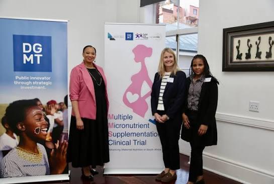 Three women standing beside a banner reading “Multiple Micronutrient Supplementation Clinical Trial. Enhancing Maternal Nutrition in South Africa.”