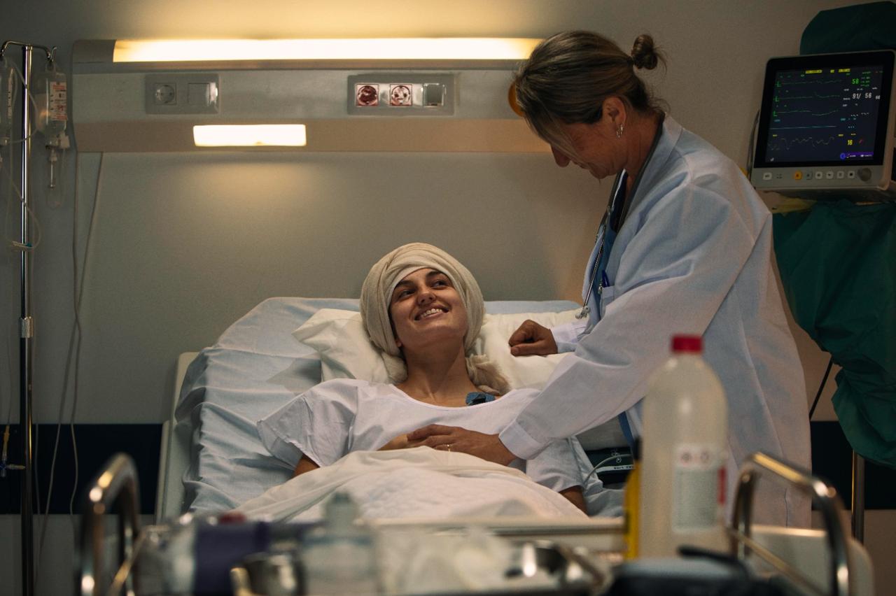 A doctor leaning over a smiling patient in a headscarf in a hospital bed, a vital-signs monitor beside them.