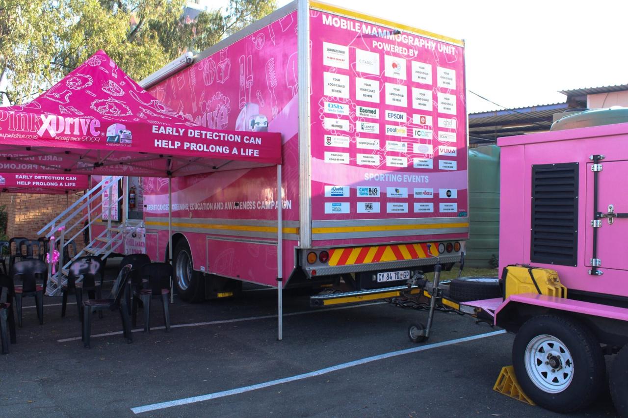 A pink PinkDrive mobile mammography unit parked with its awning up for a breast screening day, its side reading “Early detection can help prolong a life”.