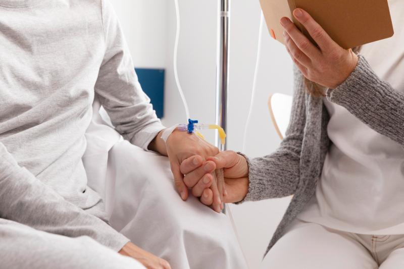 A visitor holding the hand of a patient who has a drip taped to her wrist in a hospital bed.