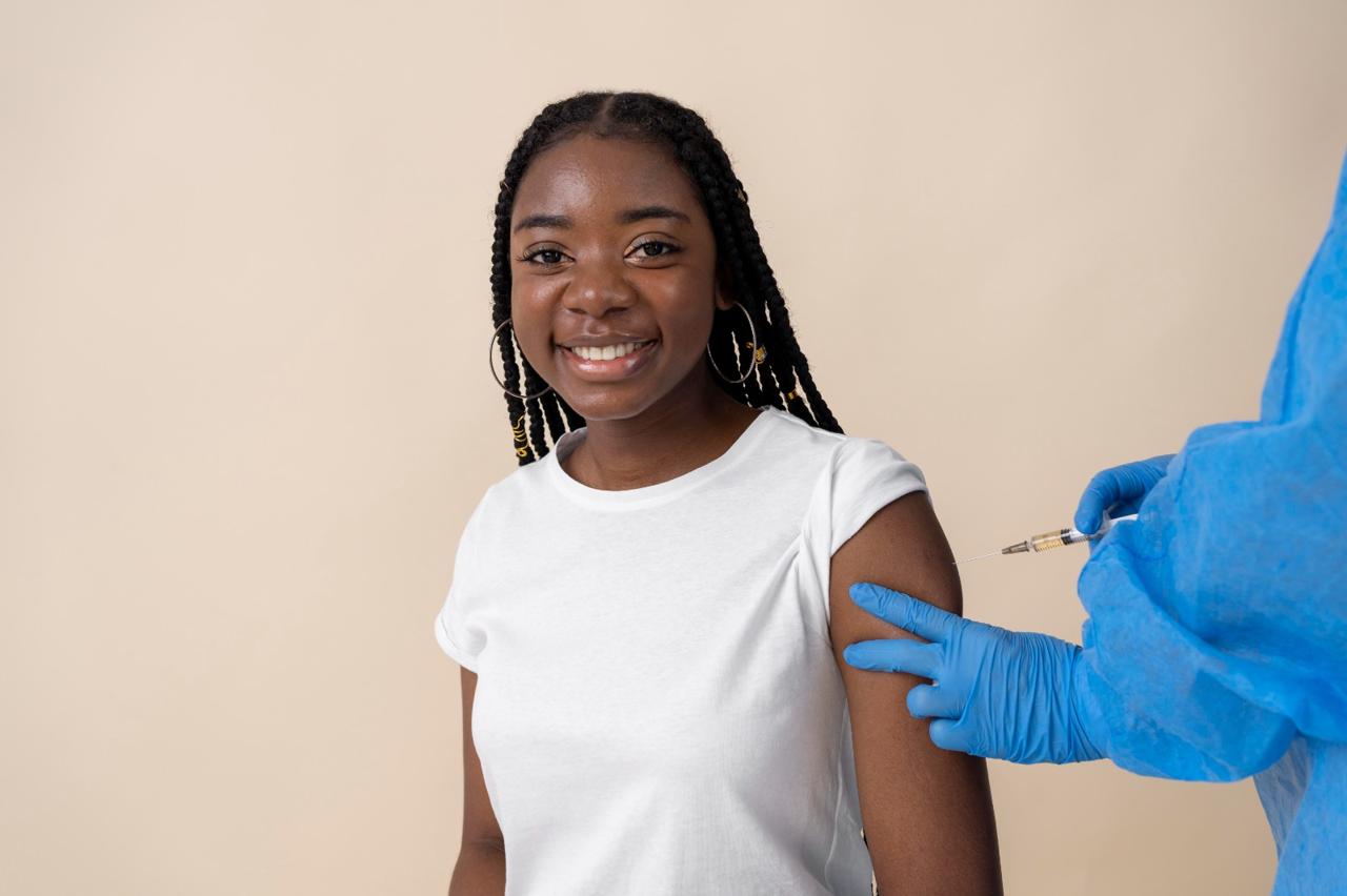 A smiling young woman in a white T-shirt receiving an injection in her upper arm from a gloved health worker.