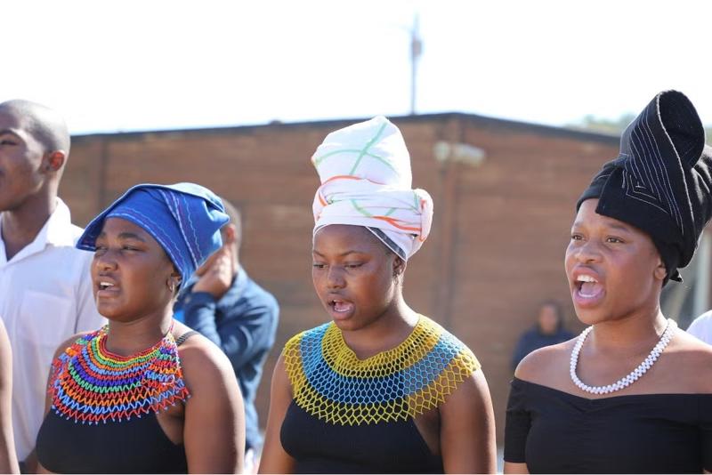 Three young women in beaded Xhosa collars and headwraps singing outdoors at a community event.