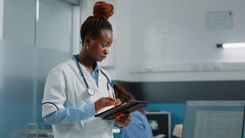 A doctor in a white coat reading a patient list on a tablet in a clinic consulting room.