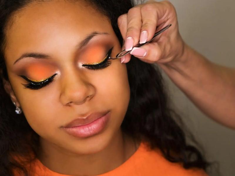 A close-up of a young woman’s closed eye as a technician applies a lash strip with tweezers.
